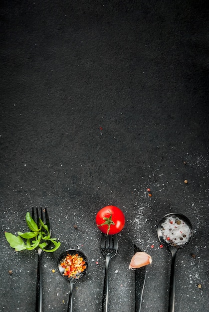 Foto hintergrund kochen. eine reihe von schwarzen geschirr - gabeln, messer und löffel mit frischen kräutern (basilikum, rucola), tomaten, knoblauch und gewürzen. auf schwarzem steintisch. draufsichtkopienraumvertikale