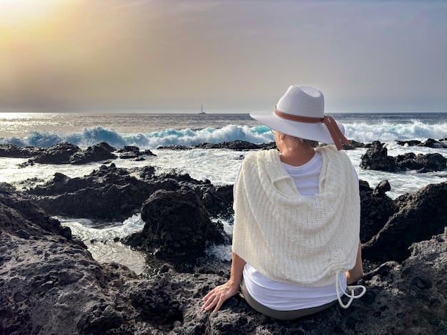 Hintergrundansicht einer älteren Frau, die an einem felsigen Strand sitzt und die Wellen des Ozeans mit weißem Schaum beobachtet
