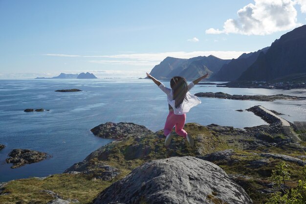 Hintergrundansicht einer Frau, die auf einem Felsen vor dem Meer gegen den Himmel springt