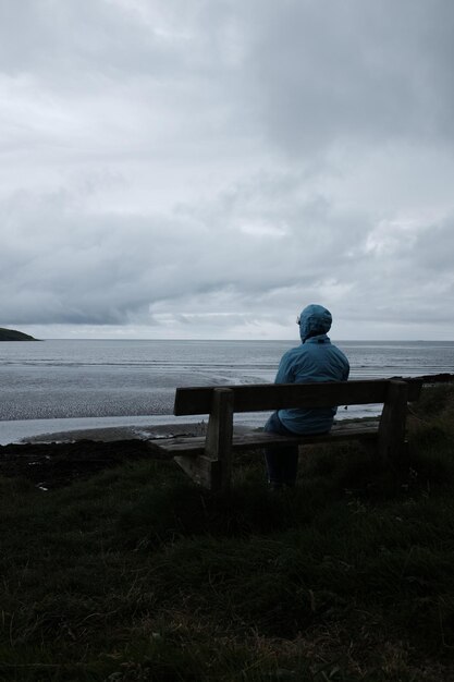 Hintergrundansicht einer Frau, die auf einer Bank am Meer gegen den Himmel sitzt