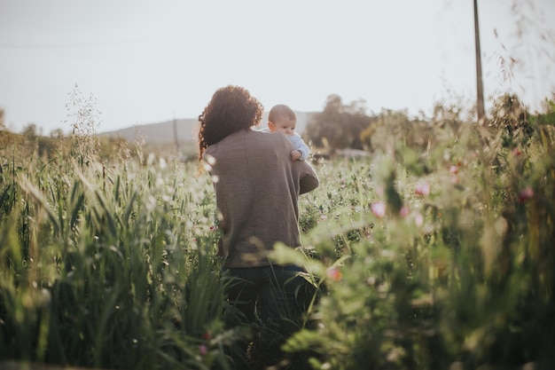 Hintergrundansicht einer Frau mit einem Baby, die auf dem Feld gegen den Himmel läuft