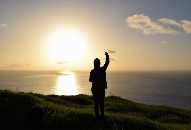 Foto hintergrundansicht einer silhouette mit erhobener hand, die während des sonnenuntergangs auf das meer und den himmel blickt