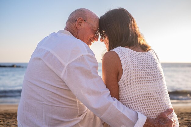 Hintergrundansicht eines älteren Paares, das sich beim Urlaub auf dem Strand umarmt und dem Meer gegenüber sitzt