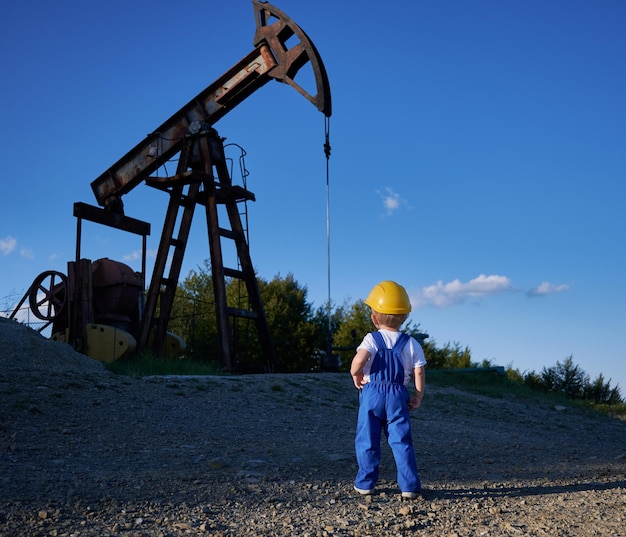 Hintergrundansicht eines intelligenten Kindes, das Geschäftsentwicklung an einer Ölstation mit Bergbauplatform und Bohrplattform studiert. Junge in gelbem Helm und blauer Uniform für Kinder, die sich den Maschinenbau ansehen