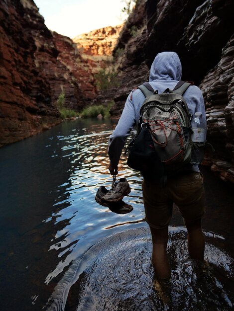 Foto hintergrundansicht eines jungen mannes mit rucksack, der in einem fluss inmitten der berge steht