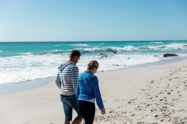 Hintergrundansicht eines kaukasischen Paares am Strand mit blauem Himmel und Meer im Hintergrund, das geht und sich die Hände hält