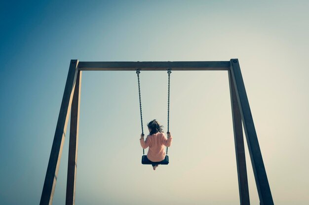 Foto hintergrundansicht eines mädchens, das auf einem spielplatz gegen einen klaren himmel auf der schaukel schwingt