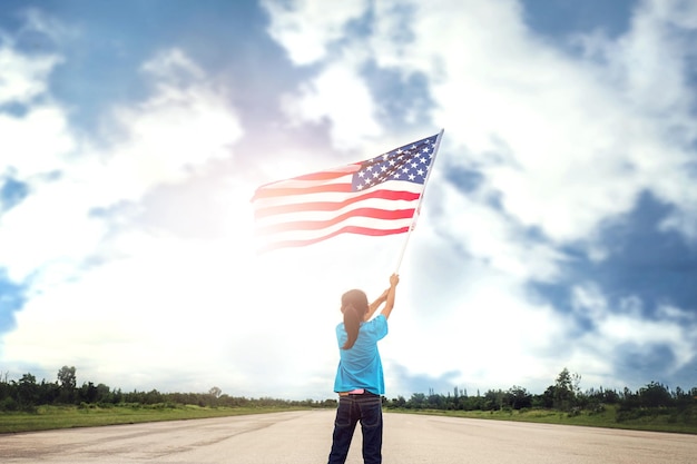 Hintergrundansicht eines Mädchens, das eine amerikanische Flagge hält, während es auf der Straße gegen den Himmel steht