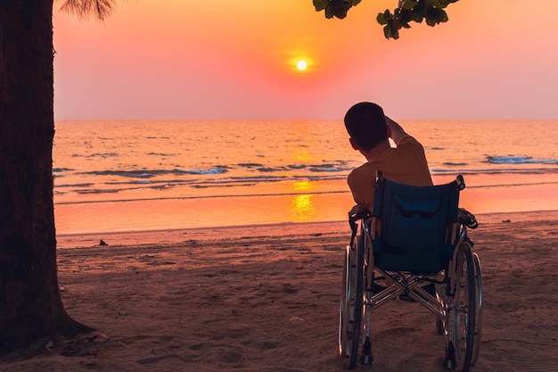 Hintergrundansicht eines Mannes, der bei Sonnenuntergang auf dem Strand im Rollstuhl sitzt