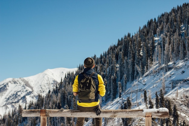 Hintergrundansicht eines Mannes, der im Winter auf einer Parkbank gegen Berge und Himmel sitzt