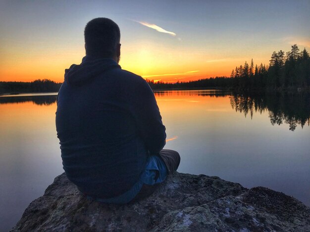 Foto hintergrundansicht eines mannes, der während des sonnenuntergangs auf einem felsen über dem see gegen den himmel sitzt