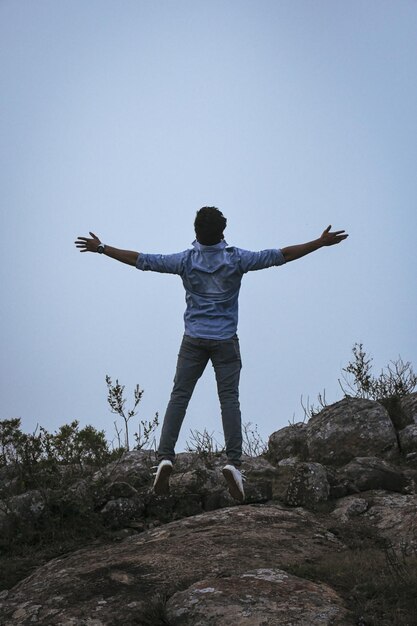 Hintergrundansicht eines Mannes mit ausgestreckten Armen, der auf einem Felsen gegen den Himmel springt