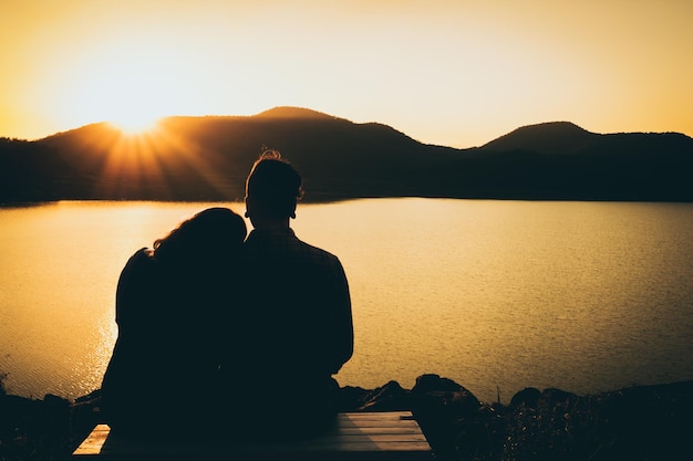 Hintergrundansicht eines Silhouette-Paares, das am See gegen den Sonnenuntergang sitzt