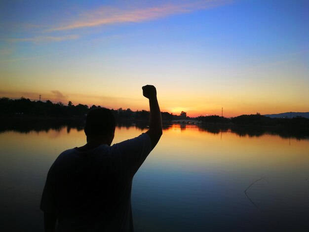 Hintergrundansicht eines Silhouettenmannes, der bei Sonnenuntergang am See gegen den Himmel steht