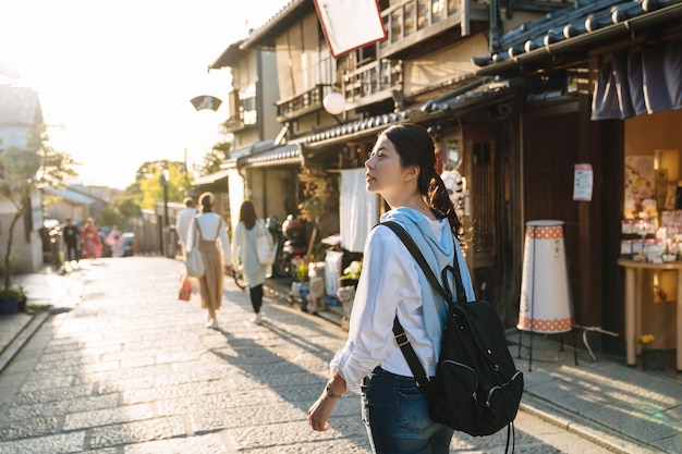 hintergrundbeleuchtetes Foto asiatische Rucksacktouristin blickt neugierig in die Ferne, während sie in der Abenddämmerung im Herbst Kyoto, Japan, die historische Straße ninen zaka hinuntergeht