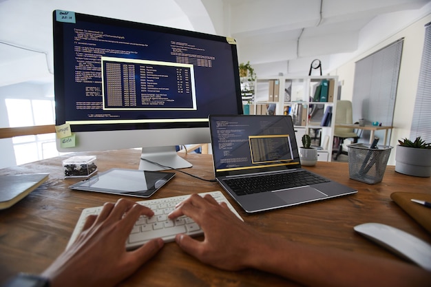 Hintergrundbild der männlichen Hände, die auf Tastatur mit schwarzem und orangeem Programmcode auf Computerbildschirm und Laptop im zeitgenössischen Büroinnenraum, Kopierraum tippen