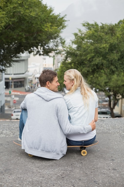 Hip joven pareja sentada en patineta sonriendo el uno al otro