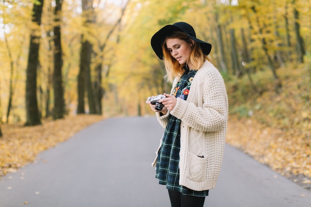 Hippiemädchen mit alter Kamera in einer gestrickten Strickjacke und in einem Hut geht Herbstpark.