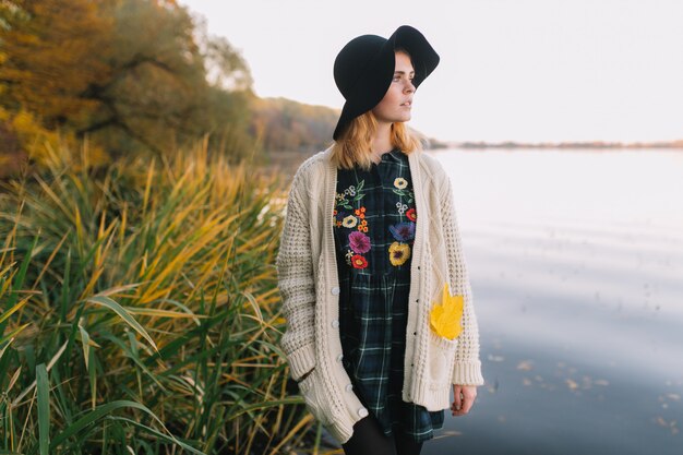 Hippiemädchen mit einem gelben Ahornblatt in einer gestrickten Strickjacke und in einem Hut geht Herbstpark.