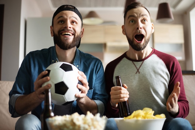 Hipster animado engraçado barbudo amigos com lanches e cervejas assistindo futebol na tv enquanto está sentado no sofá em casa. torcida gritando devido à vitória do time