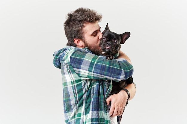 Hipster Bärtiger Typ, der einen schönen französischen Bulldoggenhund mit Liebe in seinen Armen hält und umarmt und mit ihm gegen eine weiße Wand spielt