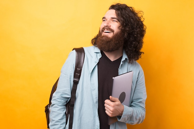 Foto hipster barbudo con cabello largo y ondulado sostiene una computadora portátil y lleva una mochila.