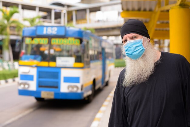 Hipster barbudo maduro sério usando máscara enquanto espera no ponto de ônibus