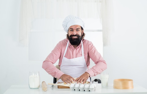 Hipster barbudo maduro sorrindo usa chapéu de chef enquanto cozinha comida na cozinha fazendo massa