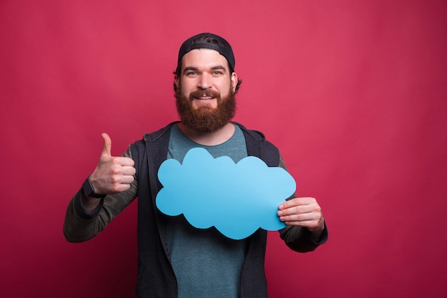 Hipster barbudo sostiene una nube azul y muestra el pulgar hacia arriba gesto
