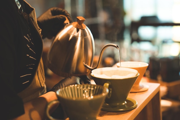 Hipster barista haciendo una bebida de café con goteo o estilo de filtro vertiendo agua caliente para preparar una bebida de cafeína de frijol negro en la tienda de café restaurante usando papel para aroma a una taza o taza