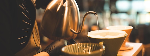 Hipster barista haciendo una bebida de café con goteo o estilo de filtro vertiendo agua caliente para preparar una bebida de cafeína de frijol negro en la tienda de café restaurante usando papel para aroma a una taza o taza