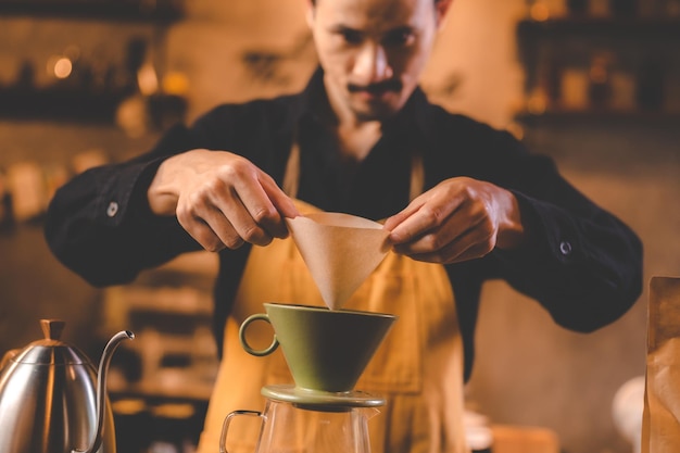 Hipster barista haciendo una bebida de café con goteo o estilo de filtro vertiendo agua caliente para preparar una bebida de cafeína de frijol negro en la tienda de café restaurante usando papel para aroma a una taza o taza