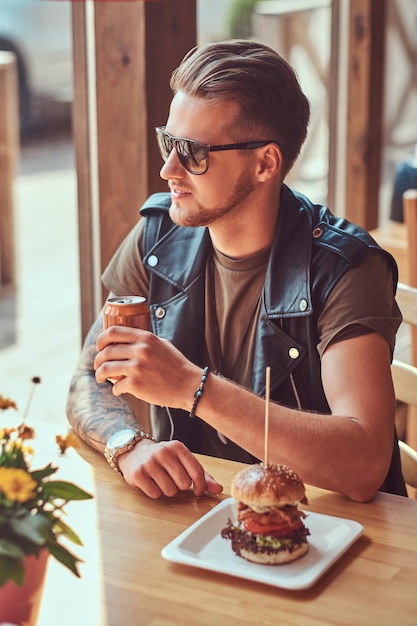 Hipster bonito com um corte de cabelo elegante e barba senta-se em uma mesa, parado para almoçar em um café à beira da estrada.