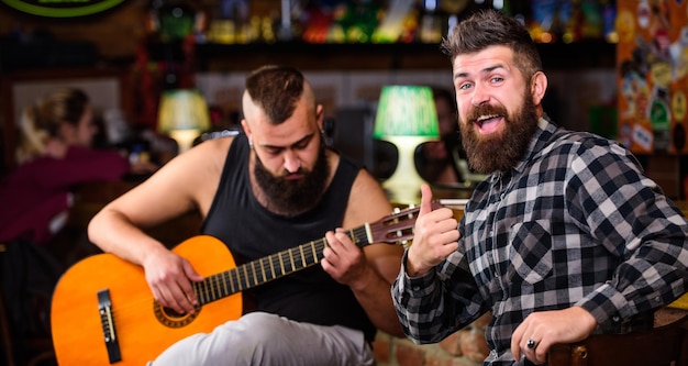 Hipster brutaler Bart mit Freund in Kneipe. Mann spielt Gitarre in der Kneipe. Fröhliche Freunde singen Gitarrenmusik. Entspannung in der Kneipe. Freunde, die sich in der Kneipe entspannen. Konzert mit Live-Musik. Akustische Leistung in der Kneipe.