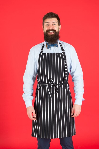 Hipster-Café-Konzept. Mann mit Bartkoch-Hipster-Schürze. Hipster-Koch kocht roten Hintergrund. Koch des bärtigen Mannes. Restaurantpersonal und Service. Gepflegter Kellner bei der Arbeit. Hipster Kellner.