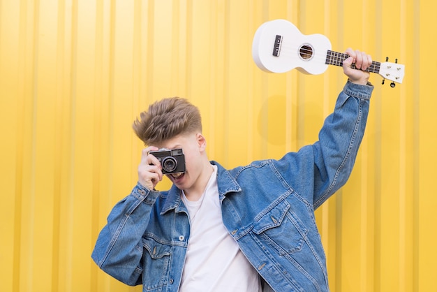 Hipster en una chaqueta de mezclilla sostiene el ukelele en sus manos y toma fotos en una vieja cámara de cine.