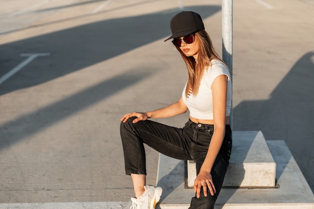 Hipster de menina elegante americana em boné preto da moda sexy top branco em jeans da moda e tênis descansar no asfalto ao ar livre em um dia ensolarado de verão. Jovem mulher urbana em roupas de juventude casual na rua.