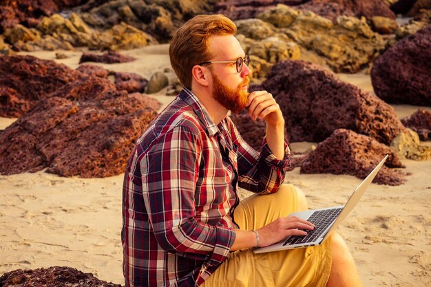 Hipster de óculos barbudo freelancer masculino com um trabalho remoto de laptop na praia por do sol. cara freelancer trabalhando perto das rochas e do mar. homem de negócios de férias em um paraíso de deserto tropical