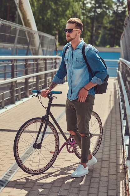 Hipster elegante bonito com um corte de cabelo elegante em óculos de sol andando com bicicleta ao ar livre.