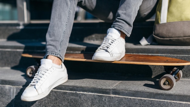 Hipster em jeans cinza e tênis branco colocou os pés no longboard Macho sentado nos degraus