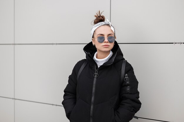 Hipster europeu elegante jovem no elegante casaco preto com um penteado moderno com uma elegante bandana posando na cidade perto de uma parede branca. roupas femininas no estilo americano.