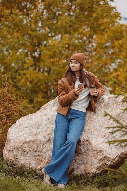 hipster femenino con taza de papel de café para llevar de pie en el parque de otoño