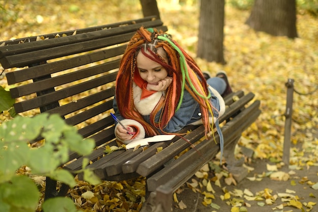 Hipster feminino escrevendo no diário no banco no parque Jovem sonhadora com longos dreadlocks deitado no banco de madeira e escrevendo notas no pequeno diário durante o fim de semana no parque outono