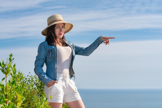 Hipster-Frau blickt in die Kamera und zeigt mit der Hand zur Seite, die auf dem Hintergrund des blauen Himmels und des Meeres steht