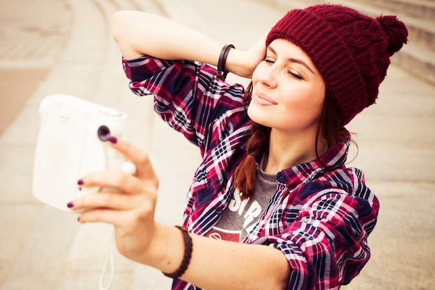 Hipster-Frau im lässigen Outfit sitzt auf Stufen in der Stadt und macht Selfie auf Sofortbildkamera