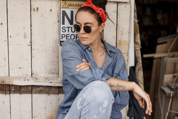 Hipster Frau in Jeans modische Kleidung mit Jeans mit einem roten Kopftuch und Sonnenbrille sitzt in der Nähe eines Vintage Holz Holz