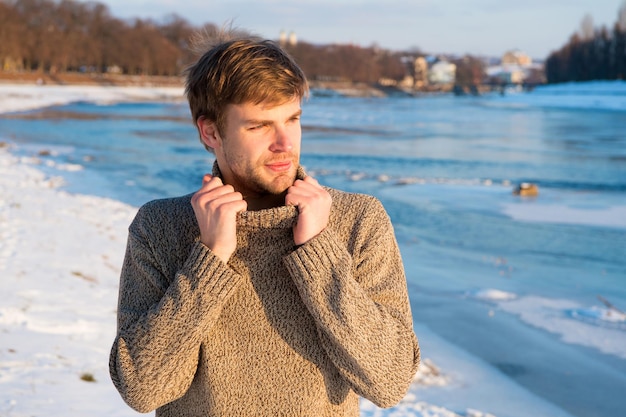 Hipster guapo sin afeitar de hombre en suéter de punto de cuello alto soporte de fondo de río congelado. Belleza del paisaje de invierno. El hombre disfruta de un día soleado de invierno. Calentamiento de ropa de invierno. Moda de invierno.