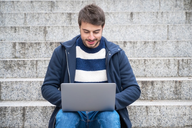 Hipster guapo empresario moderno, utilizando la computadora portátil en la ciudad, con expresión positiva.