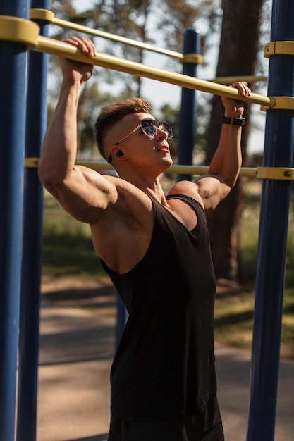 Hipster gutaussehender muskulöser Sportler mit Kopfhörern und Sonnenbrille in einem schwarzen Tanktop zieht sich an der Bar hoch und trainiert im Park einen gesunden Lebensstil