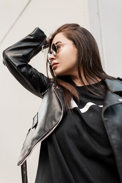 Hipster hermosa mujer fresca con gafas de sol de moda en chaqueta de cuero de moda rock y sudadera negra sobre fondo gris camina en la ciudad. Retrato urbano de una modelo femenina